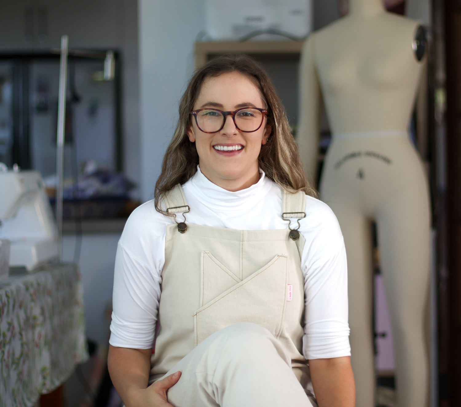 Woman wearing the yoke overalls / dungarees sitting front on to the camera smiling. Worn with a white long-sleeve underneath.