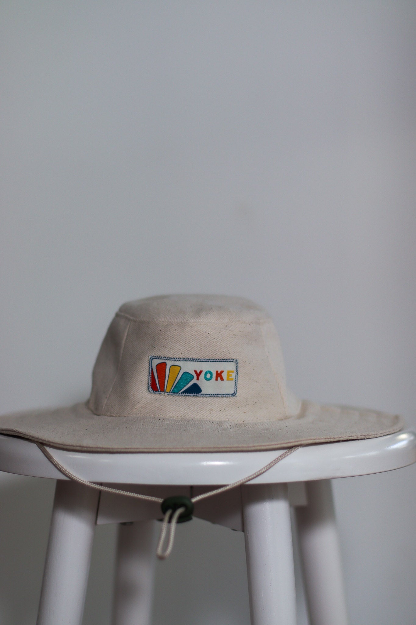 Wide brim drawstring hat. Placed on stool to display. Featuring brand label at the front. 